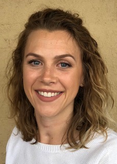 A photo image of Dr Emily Bray, a woman with long brown hair wearing a white shirt. A yellow wall can be seen in the background. 