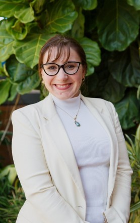 A photo image of Dr Evgeniya Zakharova-Luneva, a woman with medium length brown hair wearing a white turtle neck and jacket with a jewel necklace and black rimmed glasses. Greenery can be seen in the background. 