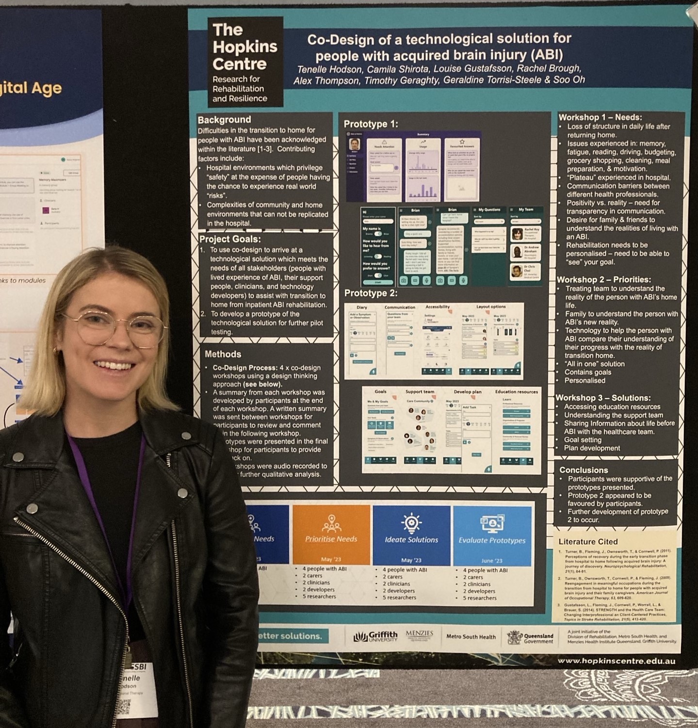 Tenelle Hodson, a woman with blonde hair and glasses wearing a black jacket, standing in front of a poster titled "Co-Design of a technological solution for people with acquired brain injury (ABI)"