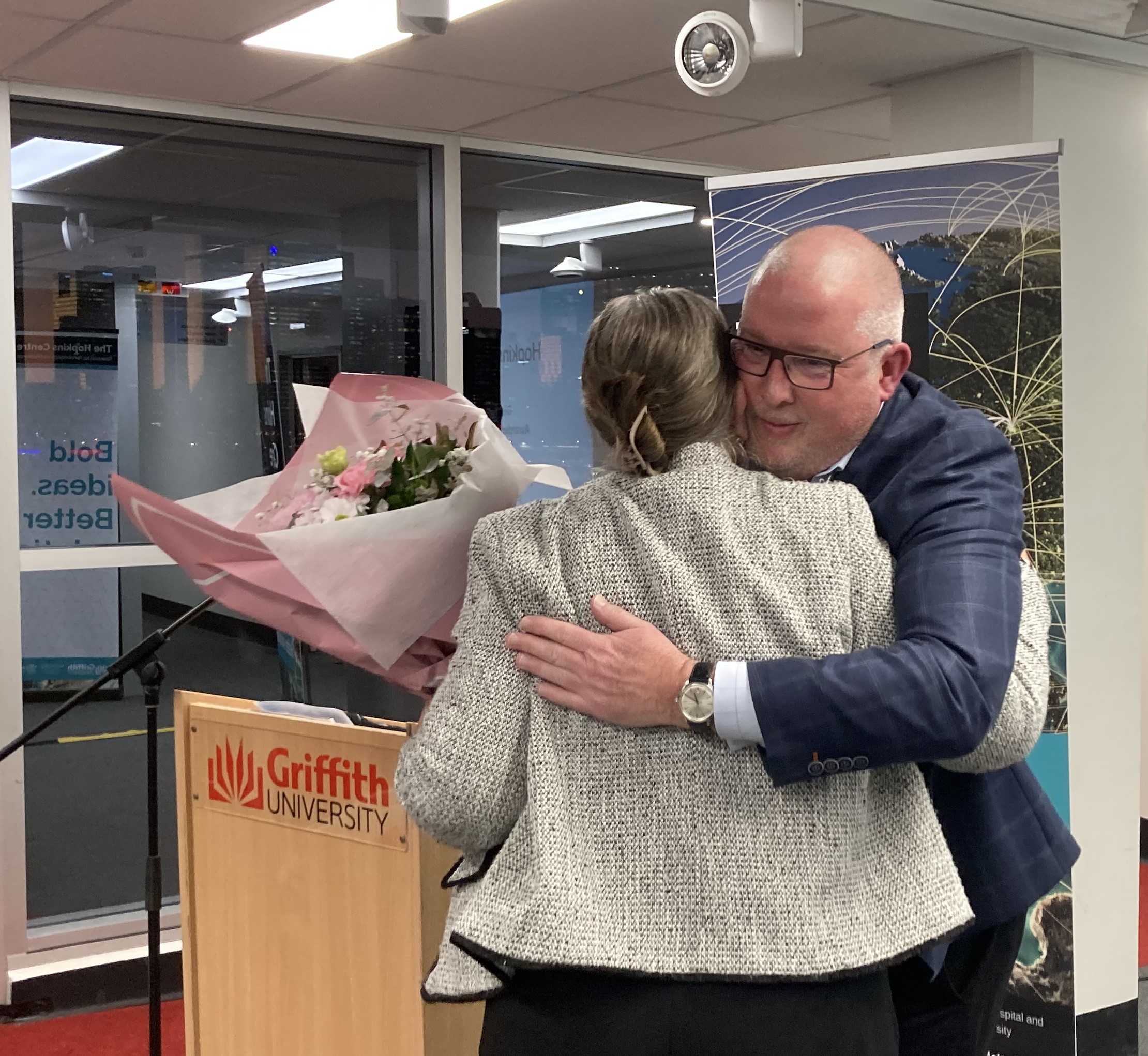 New Hopkins Centre Director, Prof. Tim Geraghty, a man with glasses wearing a blue suit jacket, and Prof. Elizabeth Kendall, a woman wearing a grey jacket with her hair clipped up, hugging as Prof Geraghty presents Prof. Kendall with a pink bunch of flowers.