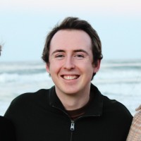 A photo image of Jayke Bennett a man with medium length black hair wearing a black sweater. Ocean waves can be seen in the background. 