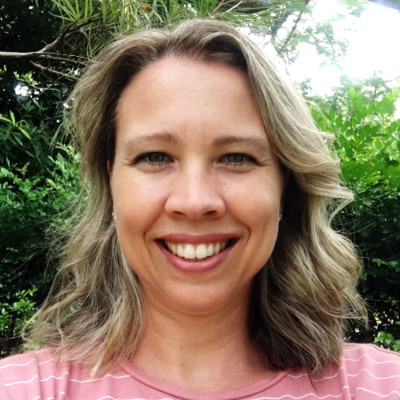 A photo image of Kristin Tynan, a woman with long blonde hair wearing a pink shirt. Greenery and the sky can be seen in the background. 