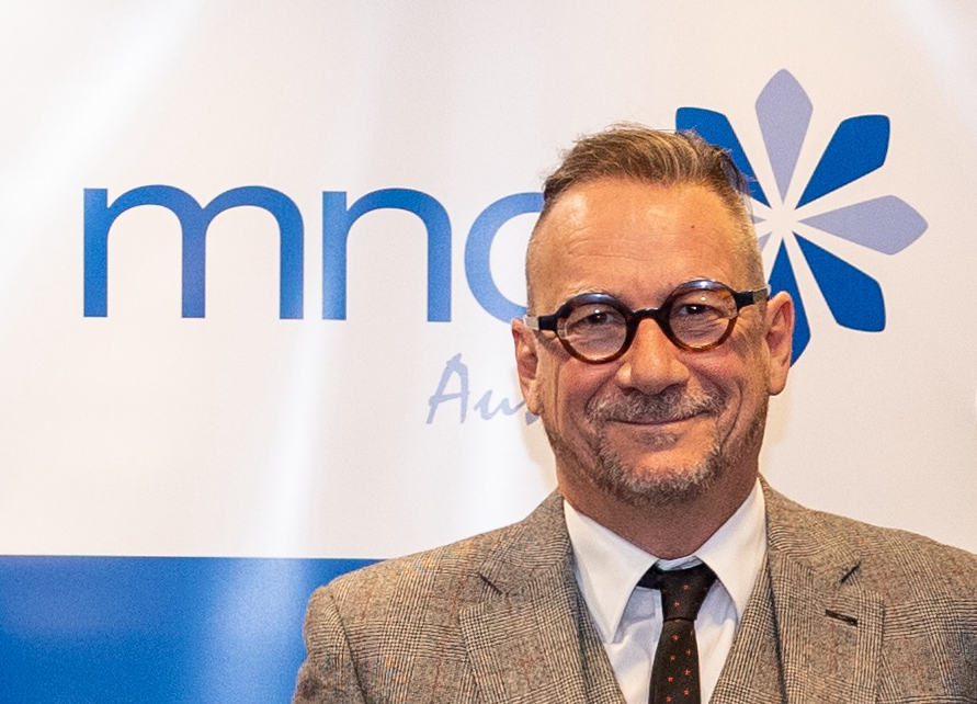 A photo image of David Berlowitz a man with short brown hair and glasses, he is dressed in a brown work jacket and white button up shirt with a dark brown tie. A presentation slide can be seen in the background.  