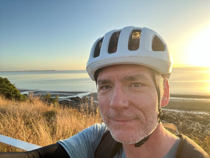 Dr Nick Aitcheson, wearing a helmet and riding gear. Behind him is the ocean and a sunset.