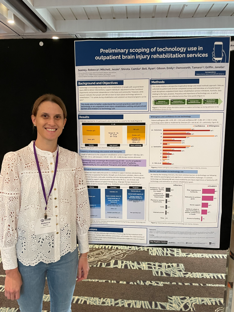 Rebecca Seeny, a woman wearing a white long sleeve top, stands in front of a poster titled "preliminary scoping of technology use in outpatient brain injury rehabilitation services"