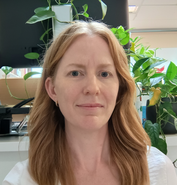 A profile photo image of Stacey James, a woman with strawberry blonde hair wearing a white top with indoor plants in the background