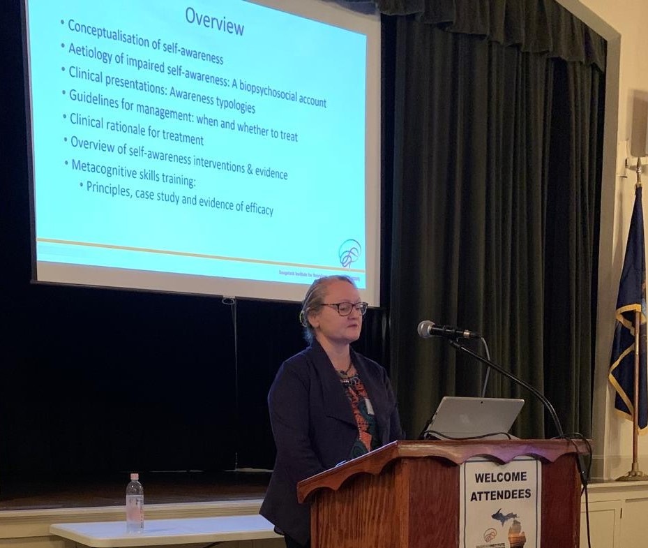 Professor Tamara Ownsworth stands behind a lectern, and in front of a large projector screen displaying the word "overview" in large bold letters, with dot points underneath.